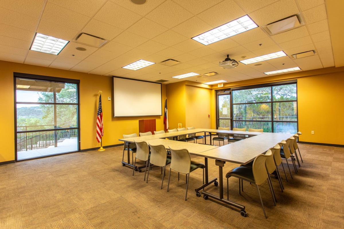 Casteel Room; 6 rectangular tables set up in a square with 18 chairs surrounding it, projector with screen dropped down, and podium